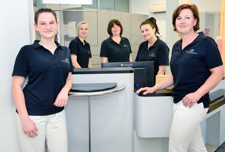 Fünf Mitarbeiterinnen einer Arztpraxis im Empfangsbereich, freundlich lächelnd in dunklen Poloshirts und heller Kleidung